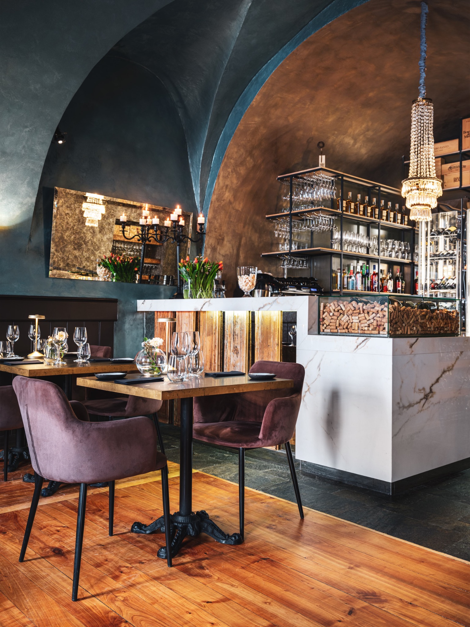 Wnętrze restauracji w Krakowie: drewniana podłoga, stoliki z aksamitnymi krzesłami, marmurowy bar z ekspozycją korków, lustro i kryształowy żyrandol.