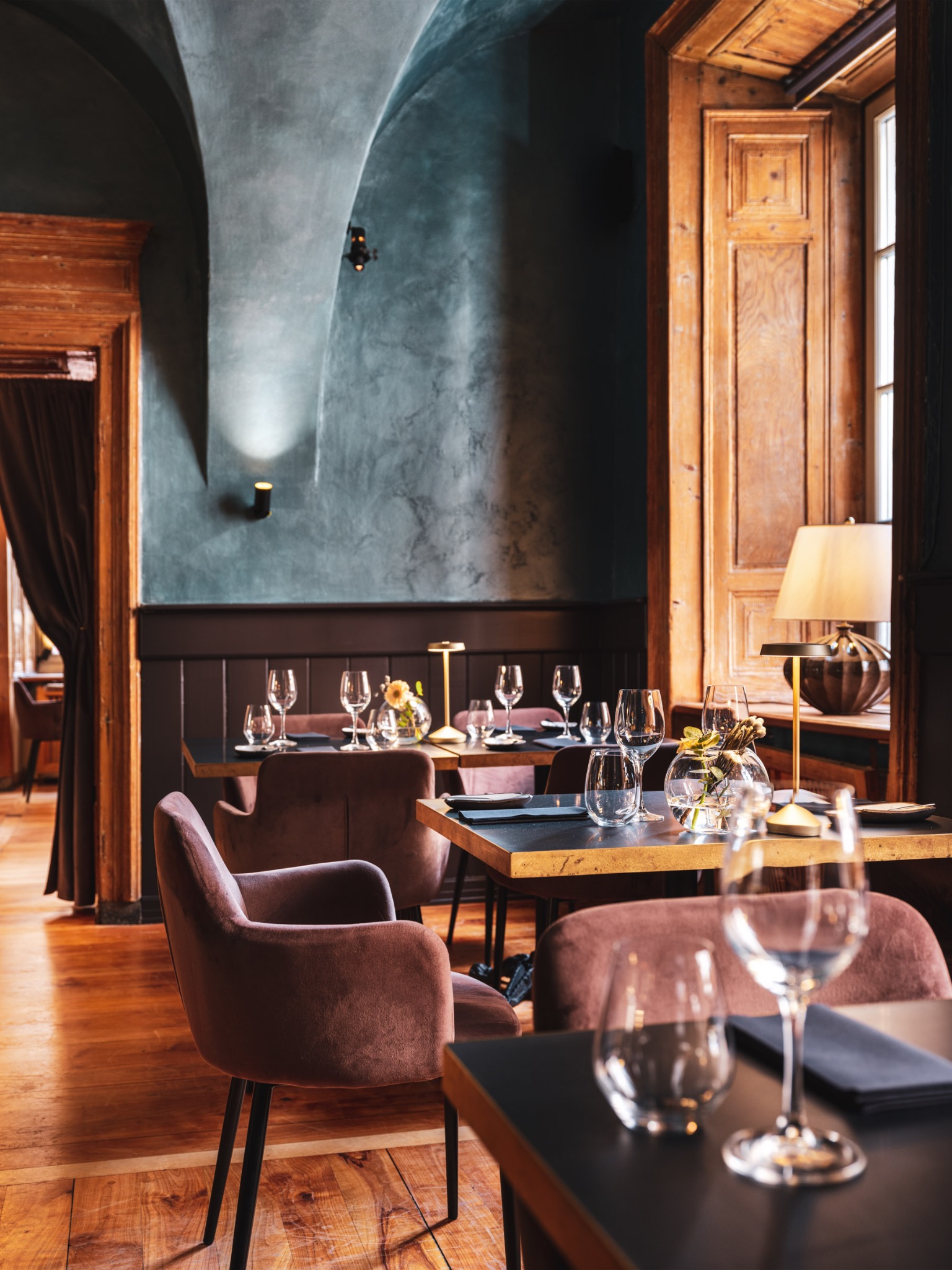 Eleganckie wnętrze restauracji z drewnianymi elementami, nakrytymi stolikami, welurowymi krzesłami i nastrojowym oświetleniem.