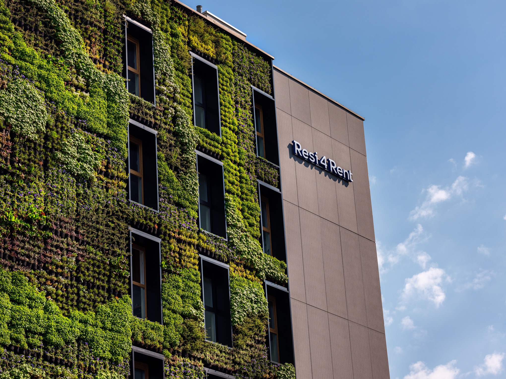 Fasada budynku Resi4Rent z pionowym ogrodem, widoczne okna i logo firmy na tle błękitnego nieba.