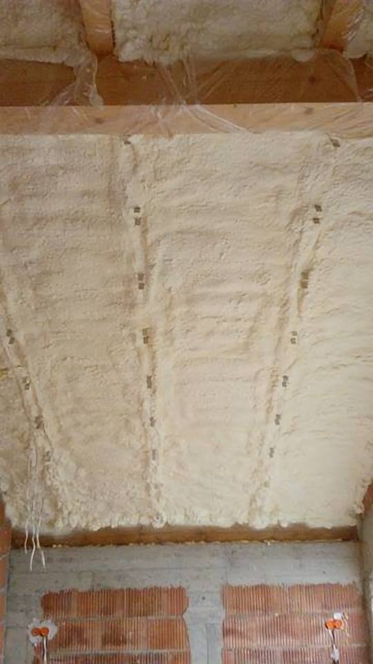 Poddasze zabezpieczone folią i pokryte grubą warstwą pianki izolacyjnej, widoczne mocowania w rzędach oraz mur z cegły dziurawki poniżej.