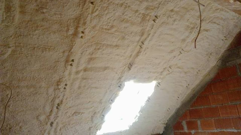 Poddasze z ociepleniem natryskowym, widoczne elementy konstrukcyjne i ceglany mur, jasne światło wpadające przez otwór.