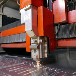 New Cut - Czerwona maszyna do cięcia laserowego precyzyjnie wycina wzór w metalowej płycie, iskry i dym unoszą się z miejsca cięcia, widoczne ślady po wcześniejszych cięciach.