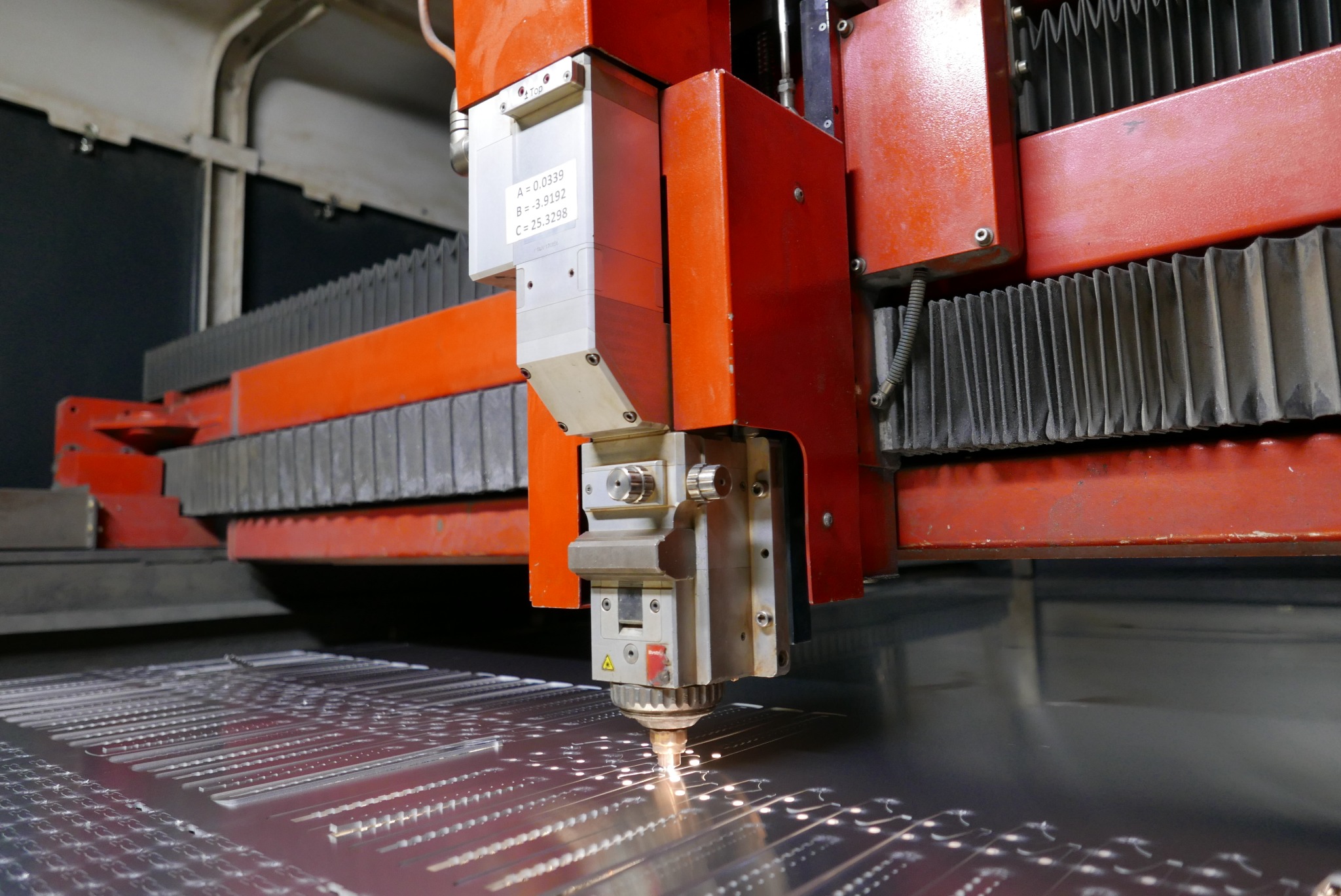 Czerwona maszyna do cięcia laserowego precyzyjnie wycina wzór w metalowej płycie, iskry i dym unoszą się z miejsca cięcia, widoczne ślady po wcześniejszych cięciach.