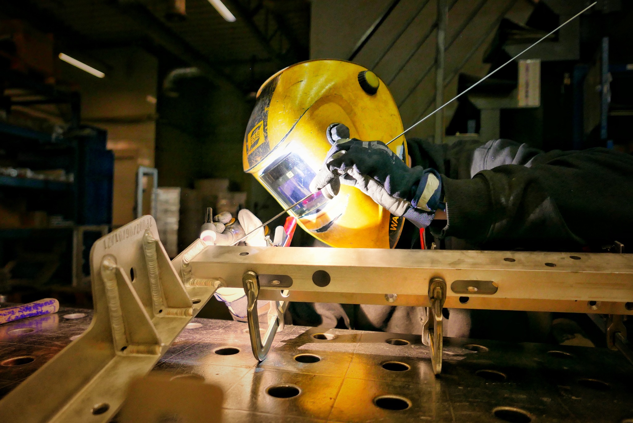 Pracownik w żółtej masce spawalniczej łączy metalowe elementy za pomocą spawarki TIG, widoczne jarzące się światło łuku elektrycznego.