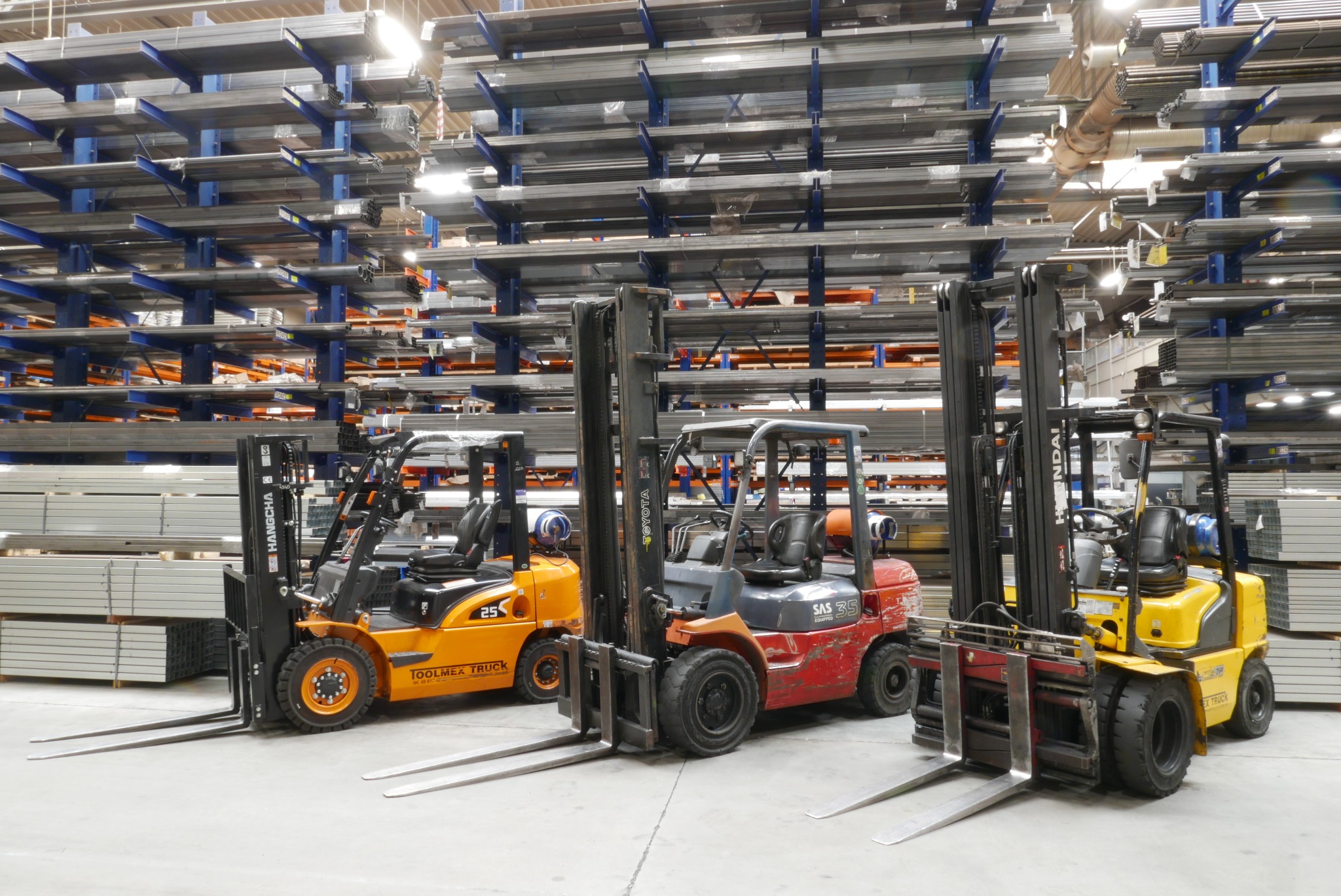 Trzy wózki widłowe różnych marek (Hangcha, Toyota, Hyundai) stojące w magazynie pełnym metalowych profili i rur składowanych na regałach.
