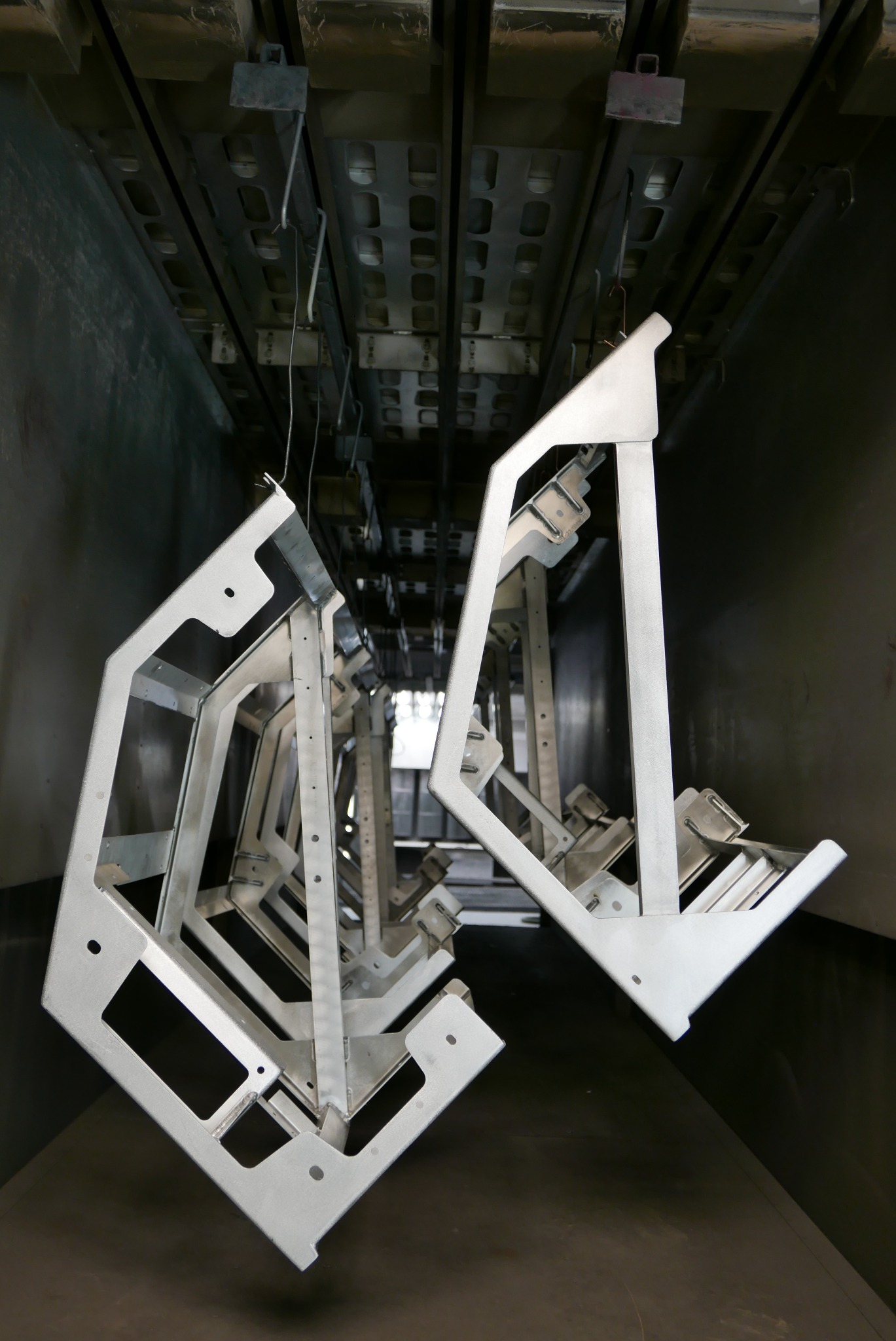 Metalowe ramy o nieregularnych kształtach, zawieszone wewnątrz lakierni proszkowej przed malowaniem, widoczne otwory montażowe i elementy konstrukcyjne.