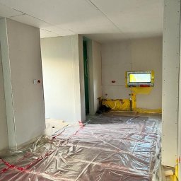 Usługi Budowlano-remontowe Tomasz Słodkiewicz - Wnętrze w trakcie prac wykończeniowych, widoczne płyty na ścianach, folia ochronna na podłodze z ogrzewaniem podłogowym i żółte zabezpieczenia okien.