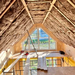 Poddasze w trakcie izolacji termicznej wełną mineralną, widoczne drewniane krokwie, okno dachowe i rusztowanie z narzędziami.