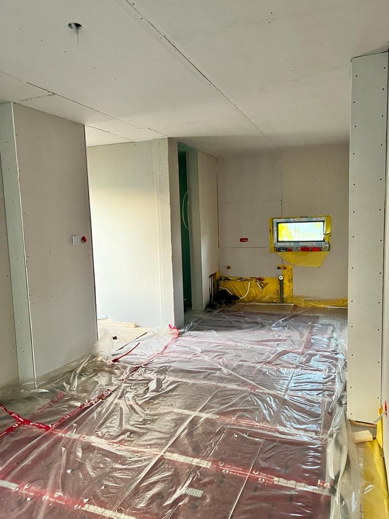 Wnętrze w trakcie prac wykończeniowych, widoczne płyty na ścianach, folia ochronna na podłodze z ogrzewaniem podłogowym i żółte zabezpieczenia okien.