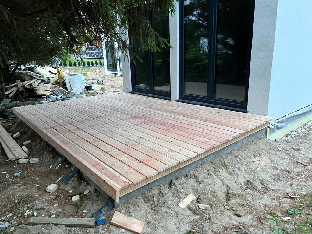 Drewniany taras w trakcie budowy przy domu z dużymi oknami, widoczne materiały budowlane i ziemia.