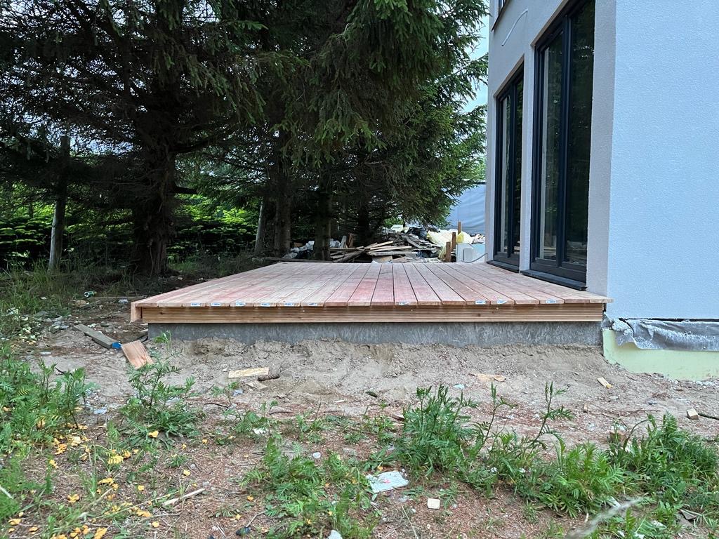 Budowa drewnianego tarasu przy nowoczesnym domu z dużymi oknami przesuwnymi, widoczne deski tarasowe i podkonstrukcja, w tle drzewa iglaste i materiały budowlane.