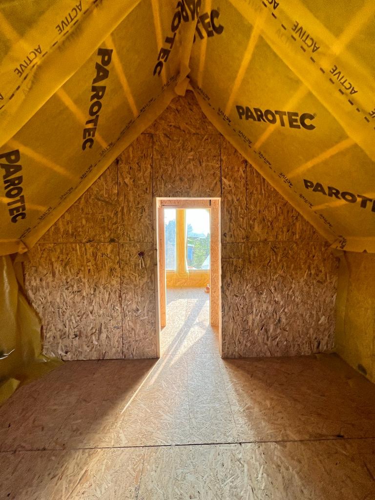 Poddasze w surowym stanie z widoczną konstrukcją dachu zabezpieczoną żółtą folią paroizolacyjną z nadrukiem 'PAROTEC', ściany obłożone płytą OSB, podłoga z desek, w tle okno z widokiem na zieleń.