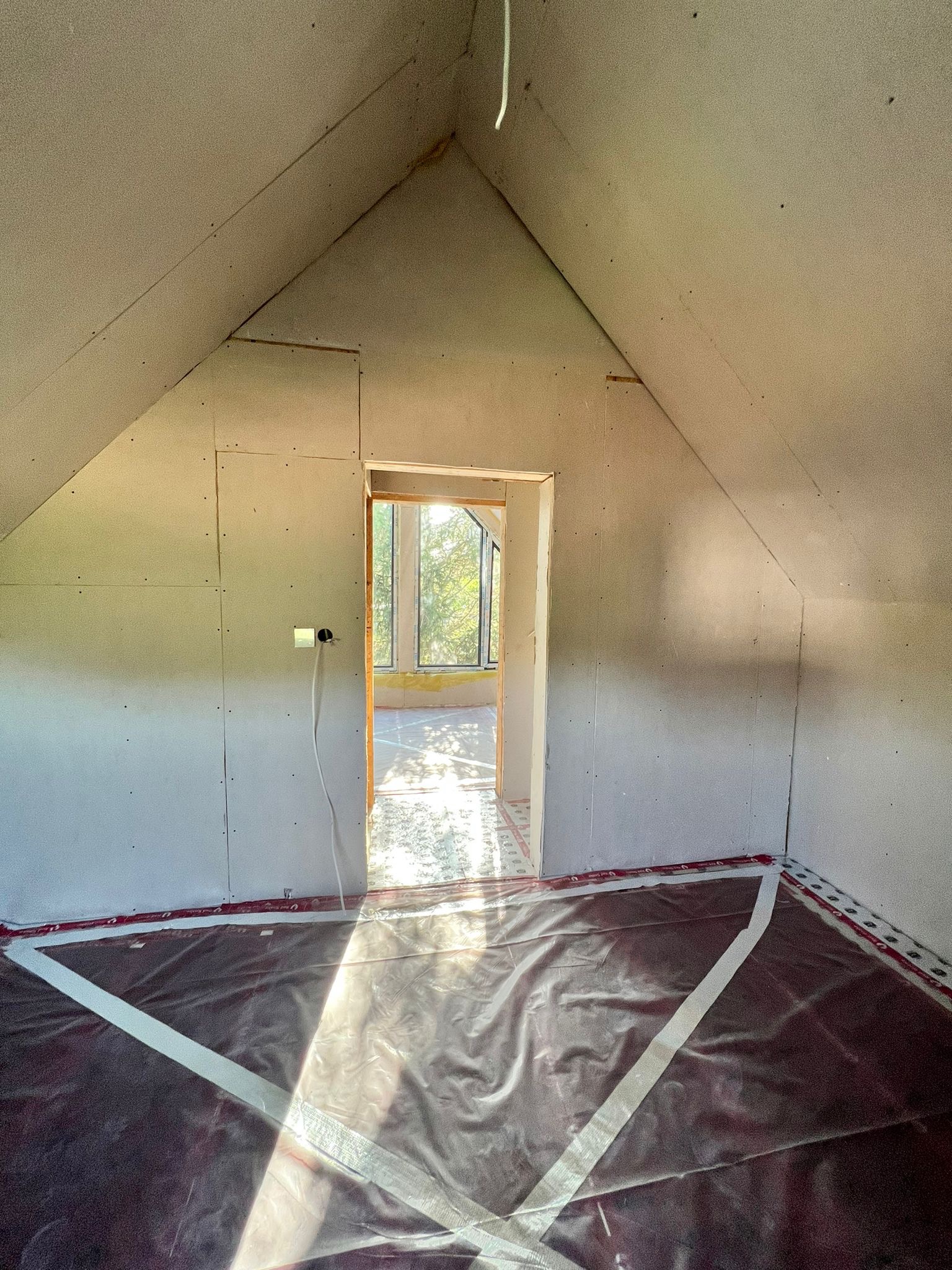 Poddasze w trakcie prac wykończeniowych: ściany obłożone płytami, zabezpieczona folią podłoga, widoczne gniazdko elektryczne i okno z widokiem na zieleń.