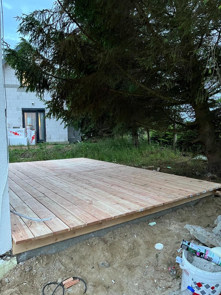 Wczesny etap budowy drewnianego tarasu przy domu w stanie surowym, widoczny fundament i deski tarasowe, w tle iglaste drzewo.