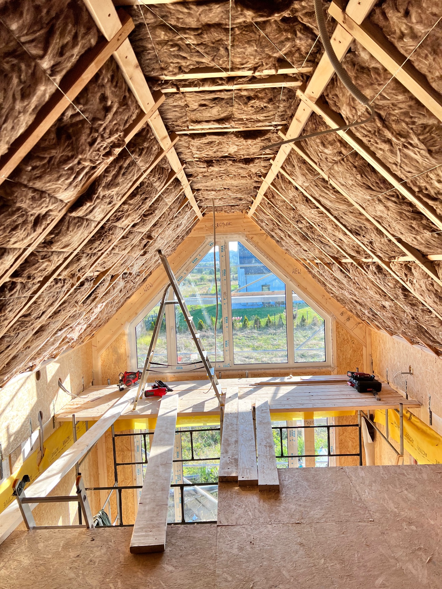 Poddasze w trakcie izolacji termicznej wełną mineralną, widoczne drewniane krokwie, okno dachowe i rusztowanie z narzędziami.