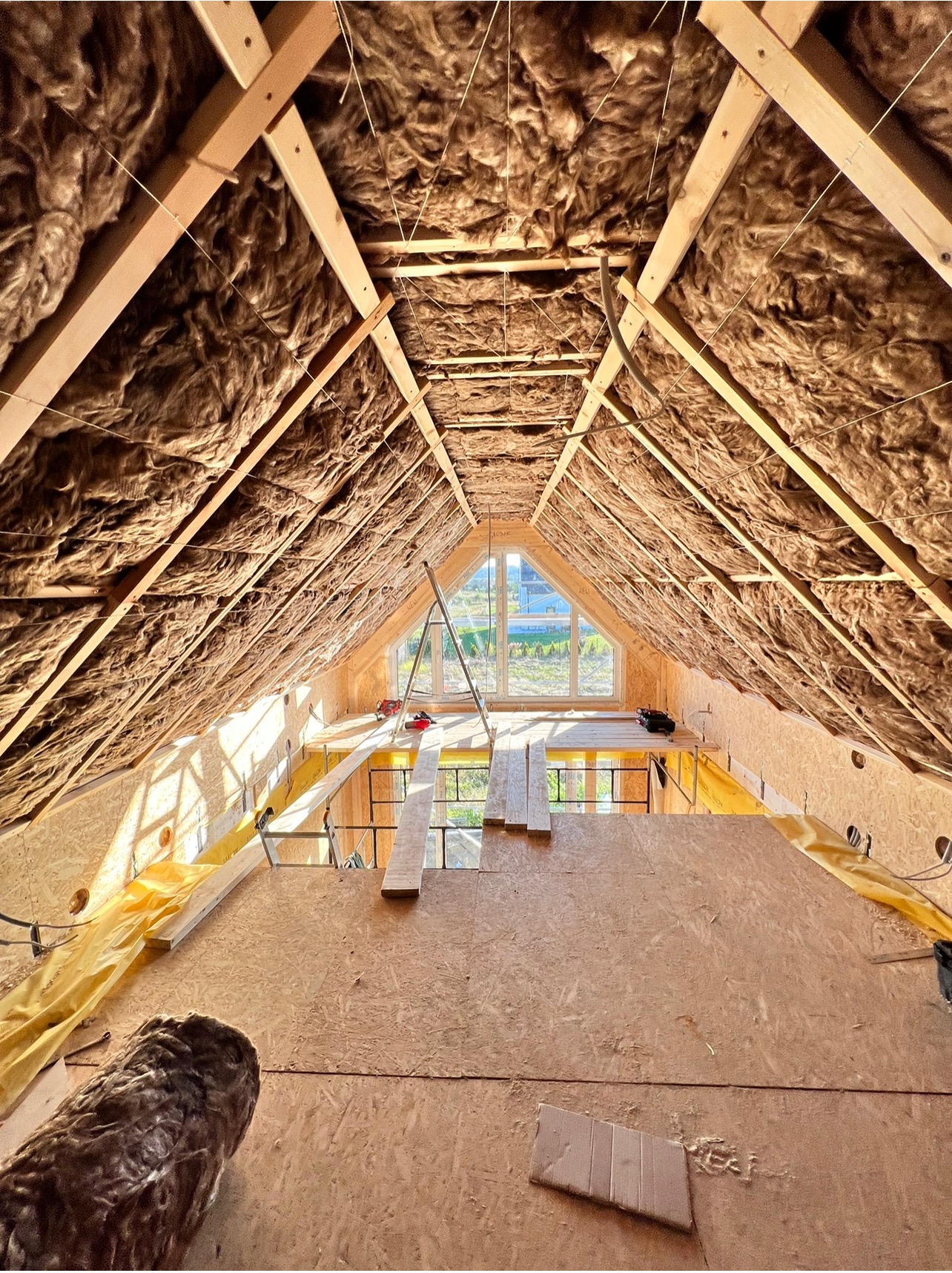 Poddasze w trakcie ocieplania wełną mineralną, widoczne drewniane krokwie i konstrukcja dachu, okno szczytowe z widokiem na zewnątrz, częściowo ułożona podłoga z płyt OSB, żółta folia ochronna...