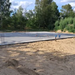 Zalana płyta fundamentowa z wystającymi prętami zbrojeniowymi, otoczona piaskiem i zielenią. Widoczne ślady opon. Słoneczny dzień.
