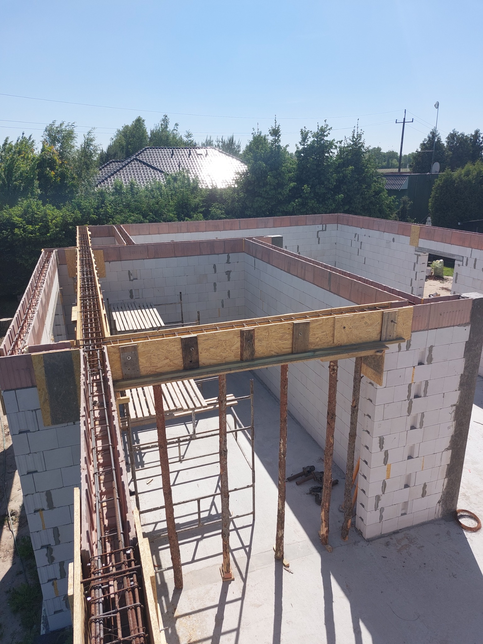 Wznoszenie ścian z bloczków betonowych z widocznym zbrojeniem w szalunkach, drewniane podpory i widok na zieleń w tle.