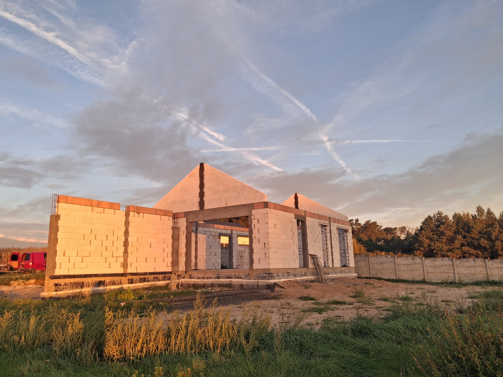 Wznoszenie ścian domu z betonu komórkowego, widoczne zbrojenia i przygotowane otwory okienne. Budowa w toku, na pierwszym planie trawa i gruz, w tle drzewa i betonowe ogrodzenie.