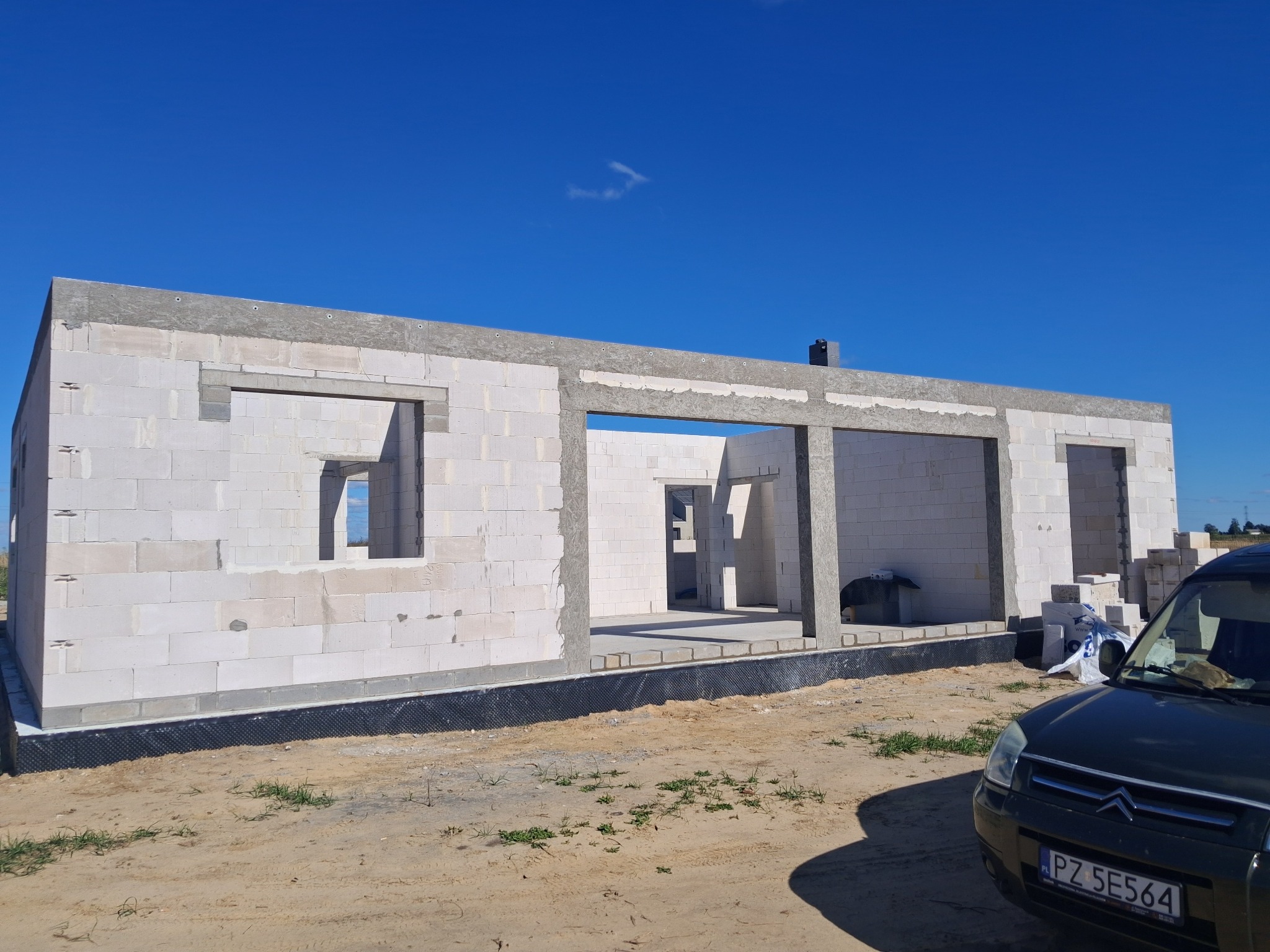 Stan surowy otwarty domu z białego bloczka, widoczne okna i drzwi, czarna izolacja fundamentów, na pierwszym planie fragment samochodu, błękitne niebo.