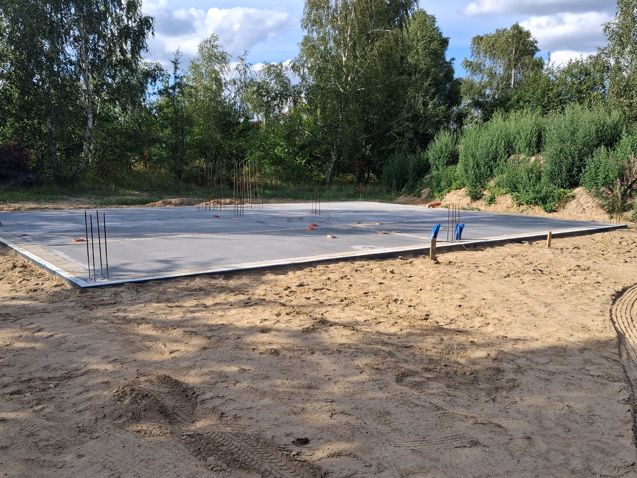 Zalana płyta fundamentowa z wystającymi prętami zbrojeniowymi, otoczona piaskiem i zielenią. Widoczne ślady opon. Słoneczny dzień.