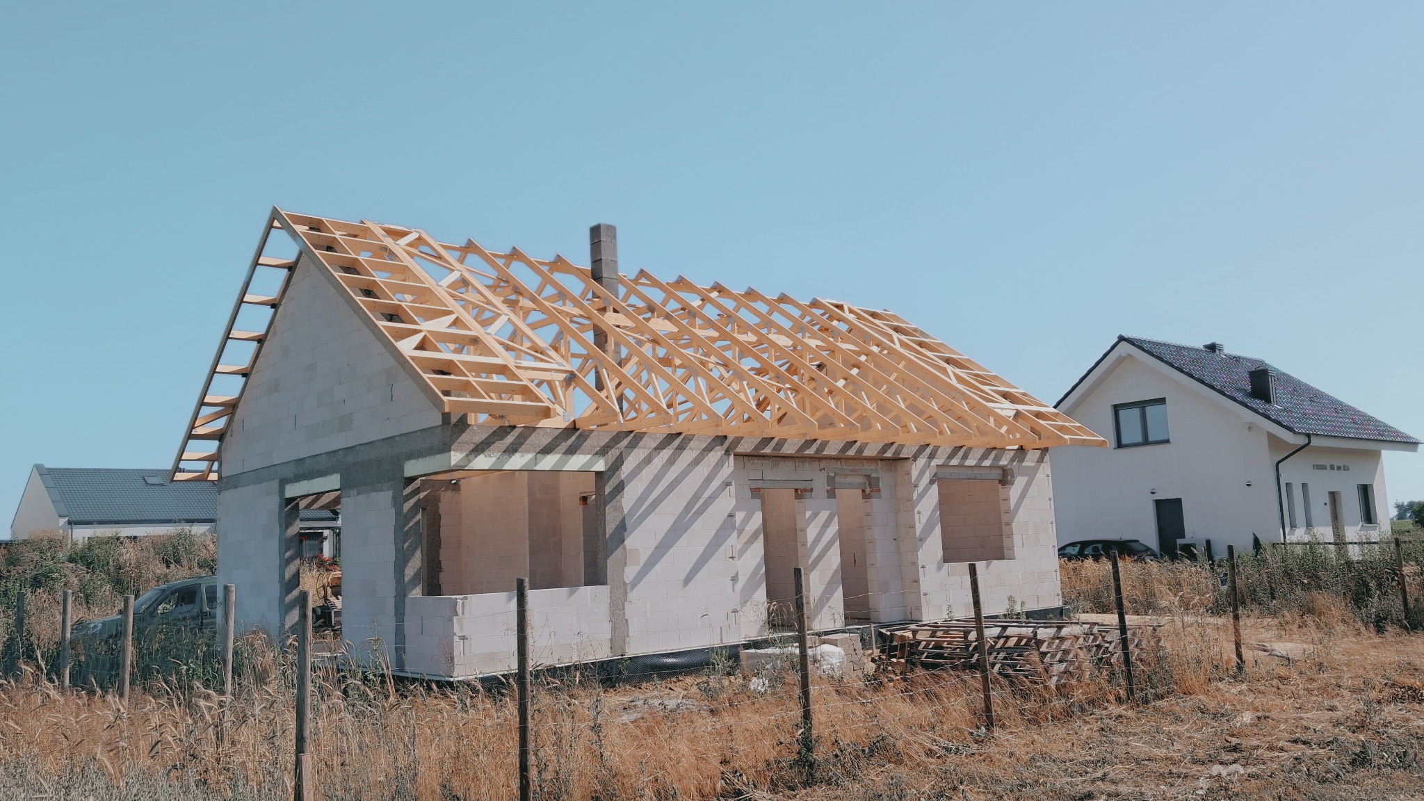 Surowy dom z bloczków z drewnianą konstrukcją dachu w trakcie budowy, na tle gotowego domu i wysokiej trawy. Widoczne palety z bloczkami.