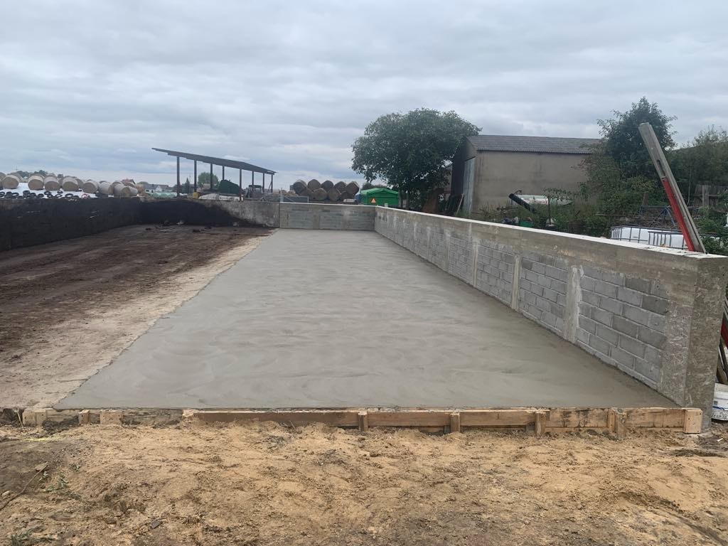 Świeżo wylany betonowy plac otoczony murem z bloczków i drewnianym szalunkiem, widok z poziomu gruntu. W tle zabudowania gospodarcze i stogi siana.