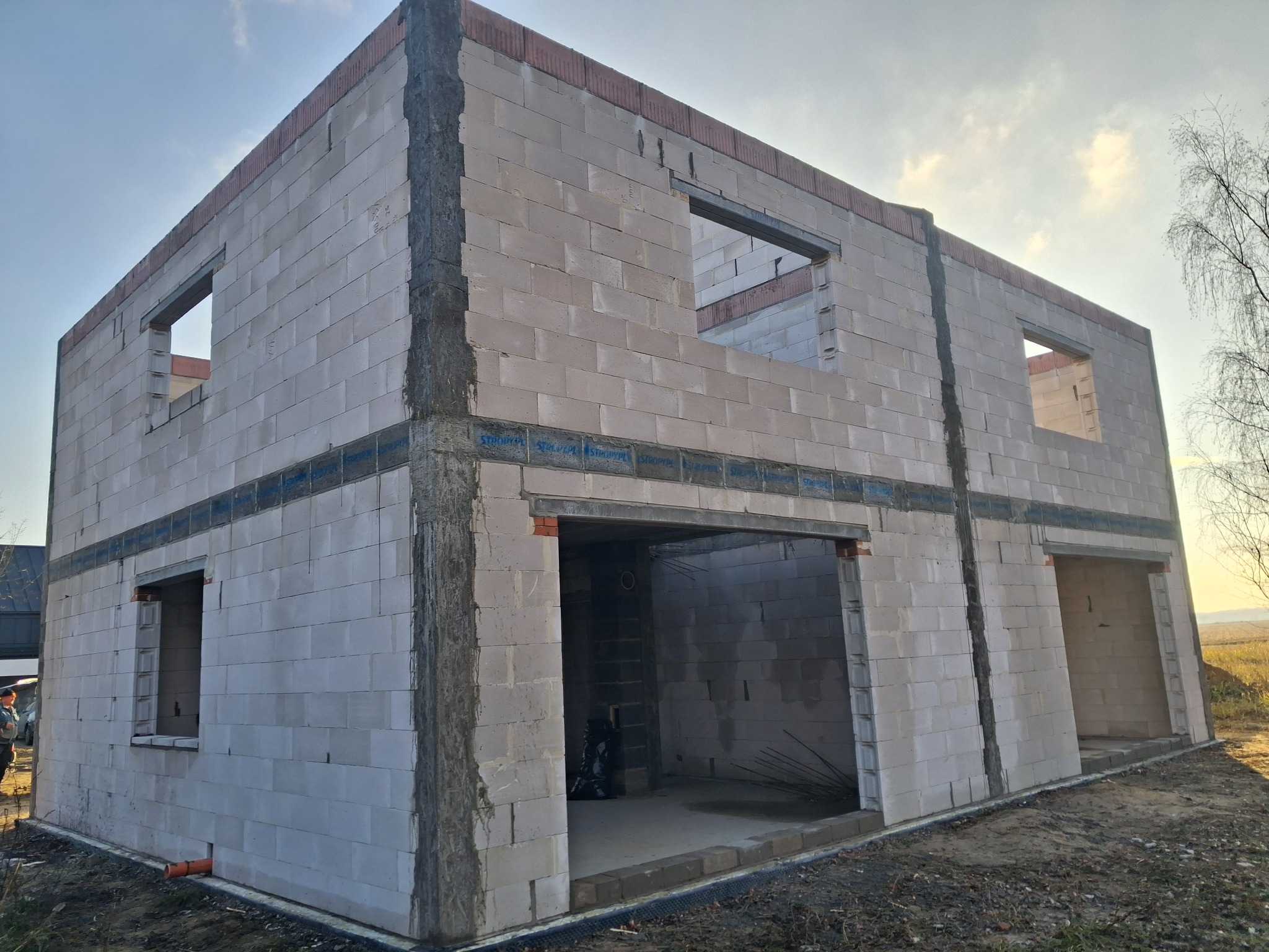 Surowy stan dwukondygnacyjnego domu z betonu komórkowego w trakcie budowy, z widocznymi otworami okiennymi i drzwiowymi, na tle błękitnego nieba i drzew.