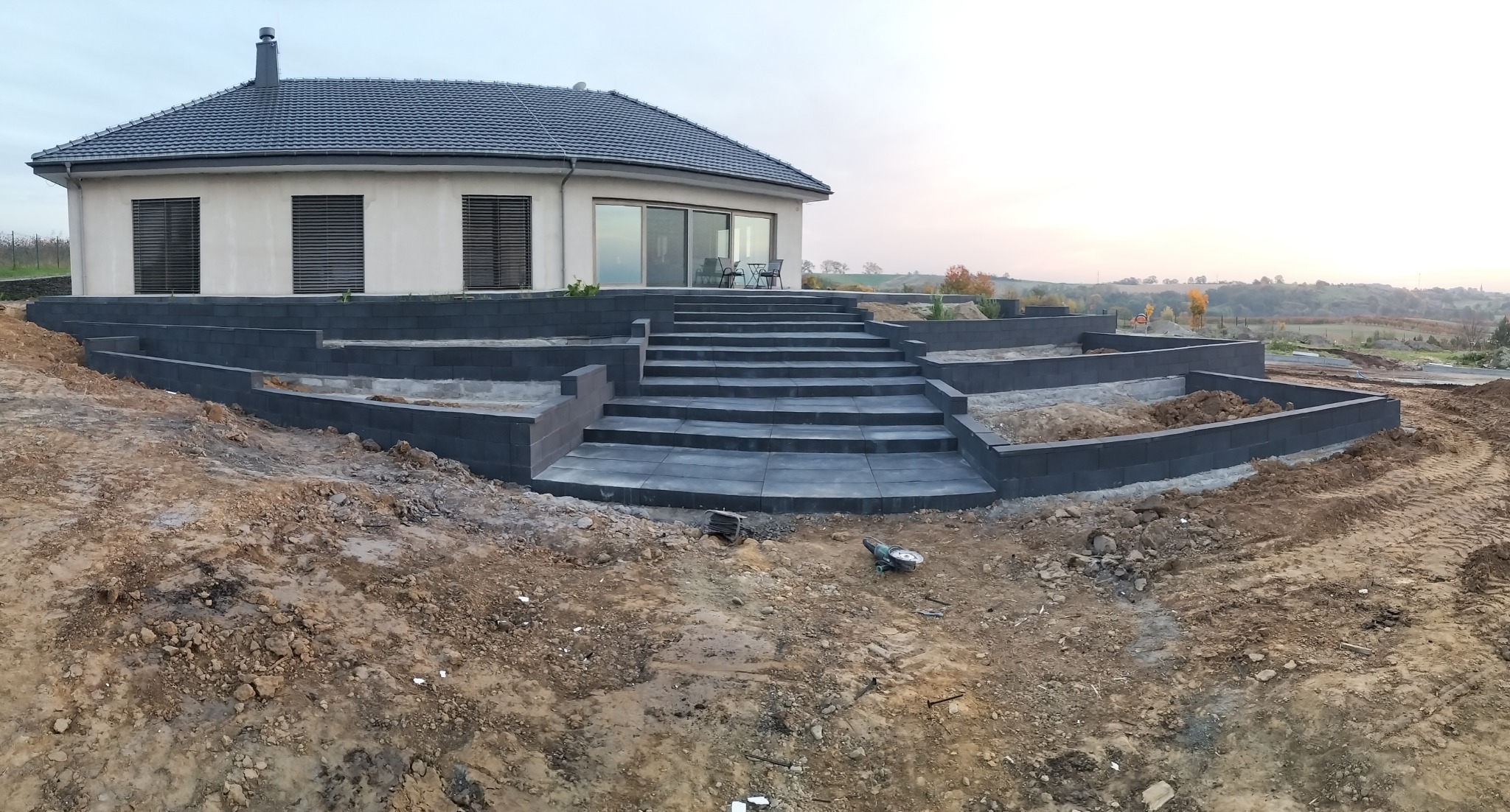 Nowoczesny dom z tarasem i schodami z kostki brukowej w trakcie budowy. Widoczne narzędzia budowlane i ziemia. Tło stanowi krajobraz wzgórz.