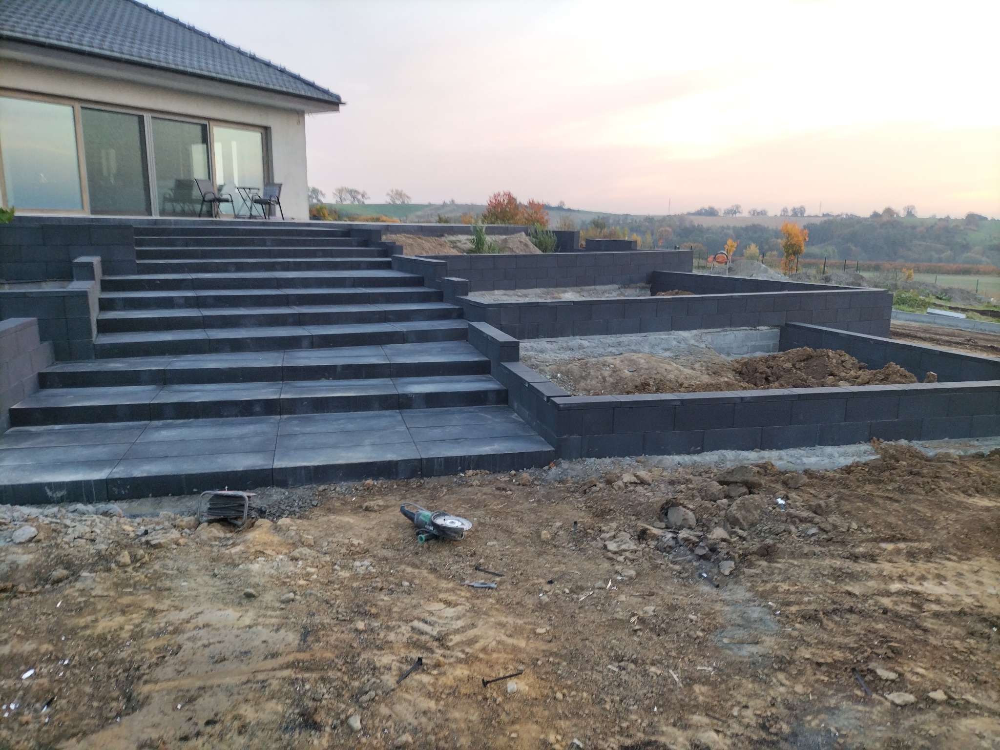 Schody i murek oporowy z szarych bloczków betonowych w ogrodzie, w tle dom i krajobraz. Na ziemi leży szlifierka kątowa. Prace w toku.
