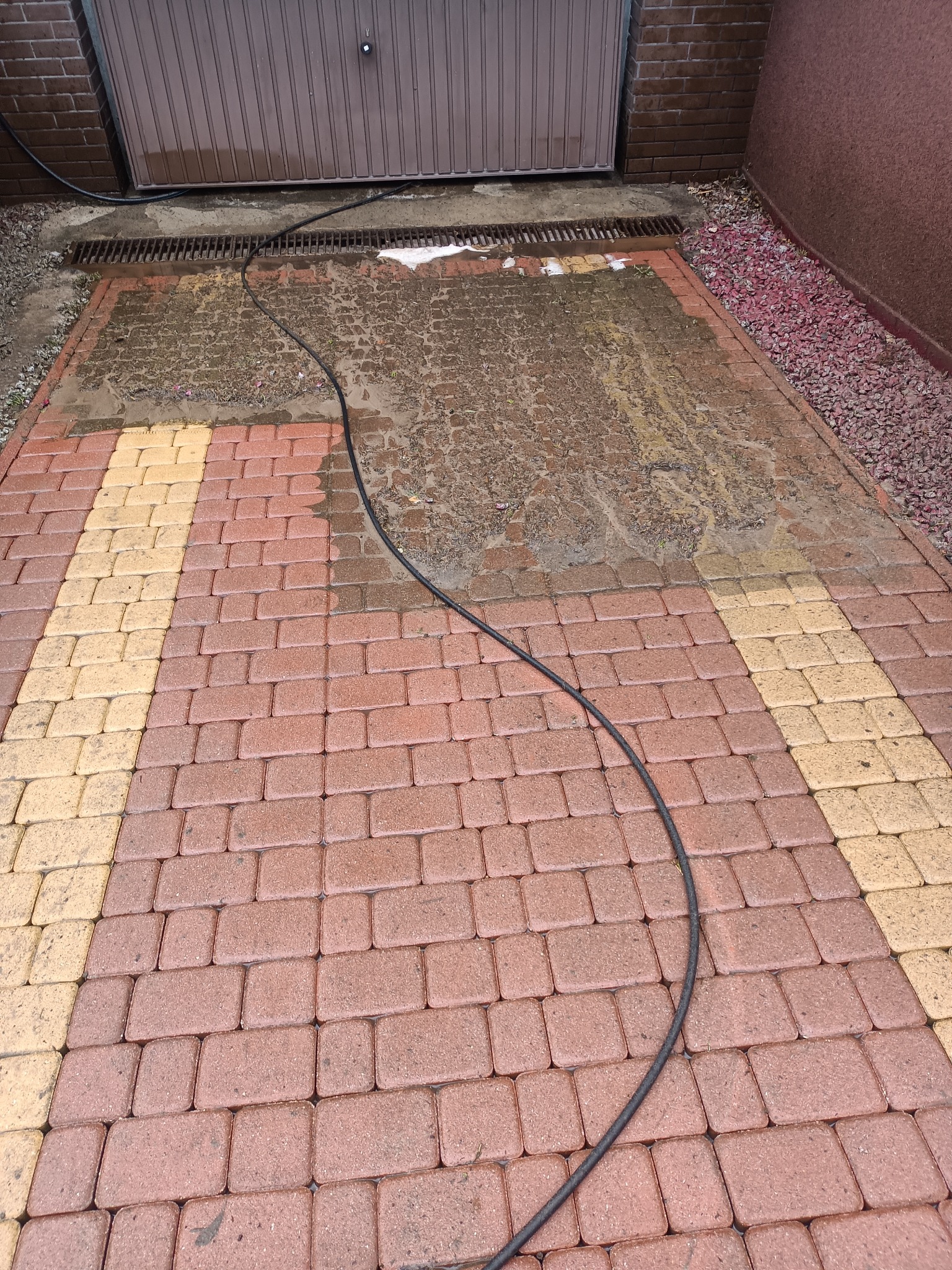 Dwukolorowa kostka brukowa częściowo umyta, z widoczną różnicą w czystości, w tle brama garażowa i wąż ciśnieniowy.