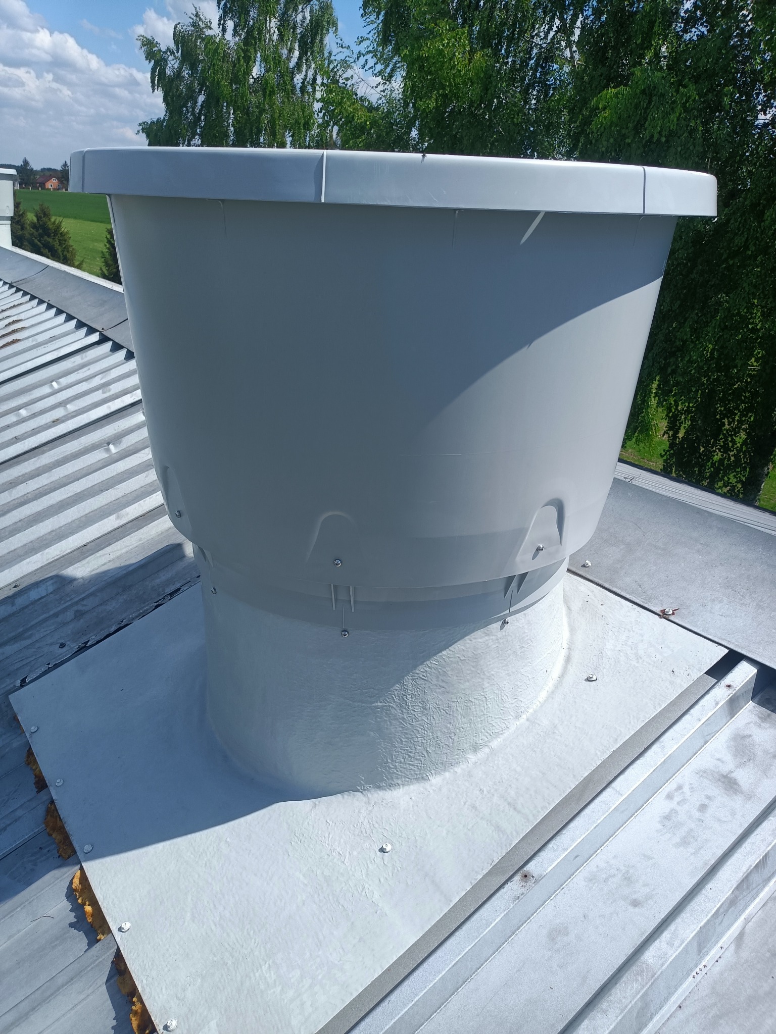 Szary komin wentylacyjny na metalowym dachu budynku, widok z góry, w tle zieleń drzew i fragment zabudowań.