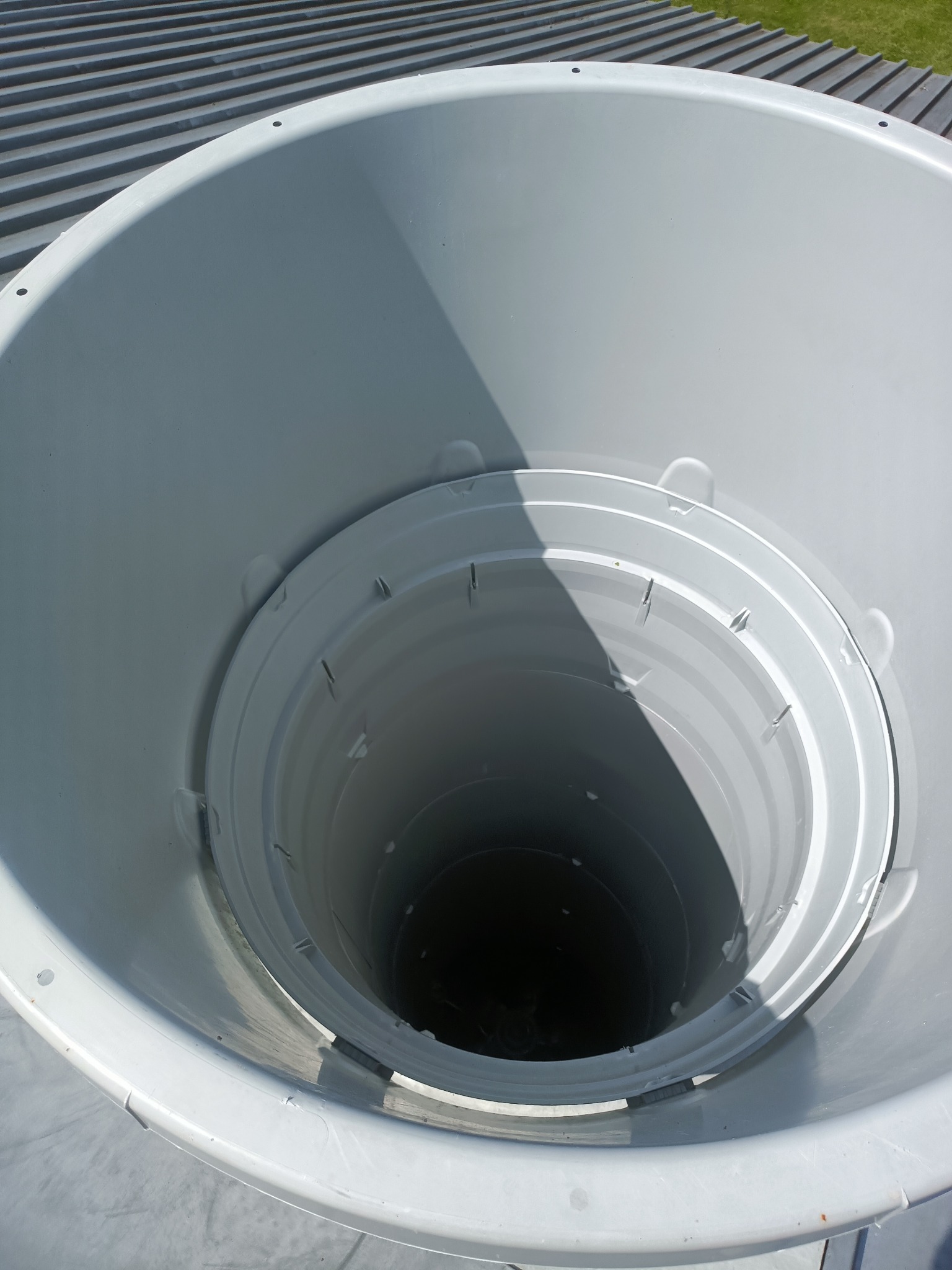 Wnętrze białego zbiornika na wodę deszczową z widocznymi elementami konstrukcyjnymi i cieniem, na tle szarego dachu i fragmentu trawnika.