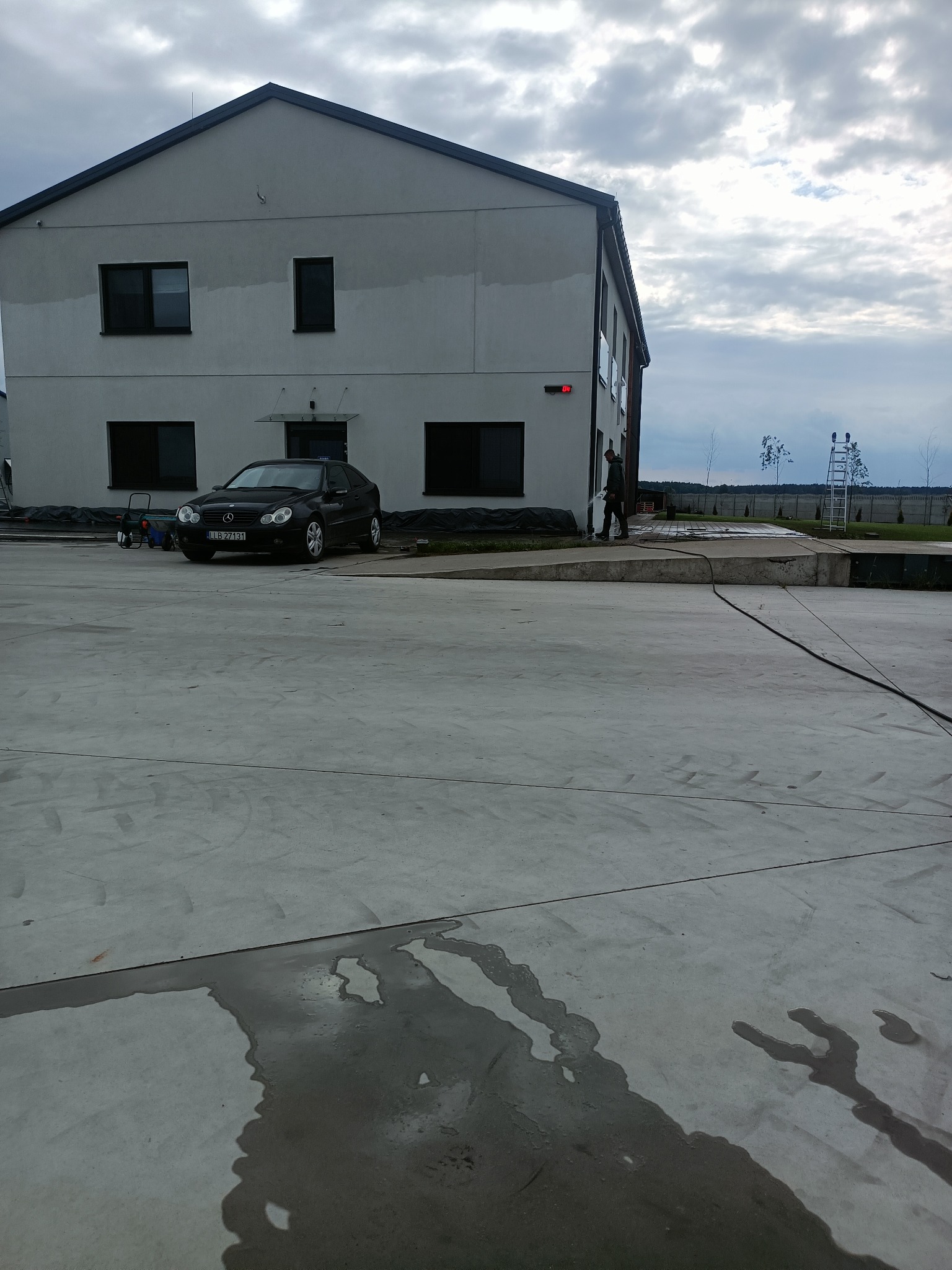 Czyszczenie elewacji budynku dwupiętrowego z czarnym samochodem zaparkowanym obok, widoczne ślady mokrej powierzchni po czyszczeniu, pracownik z lancą w prawej części kadru.