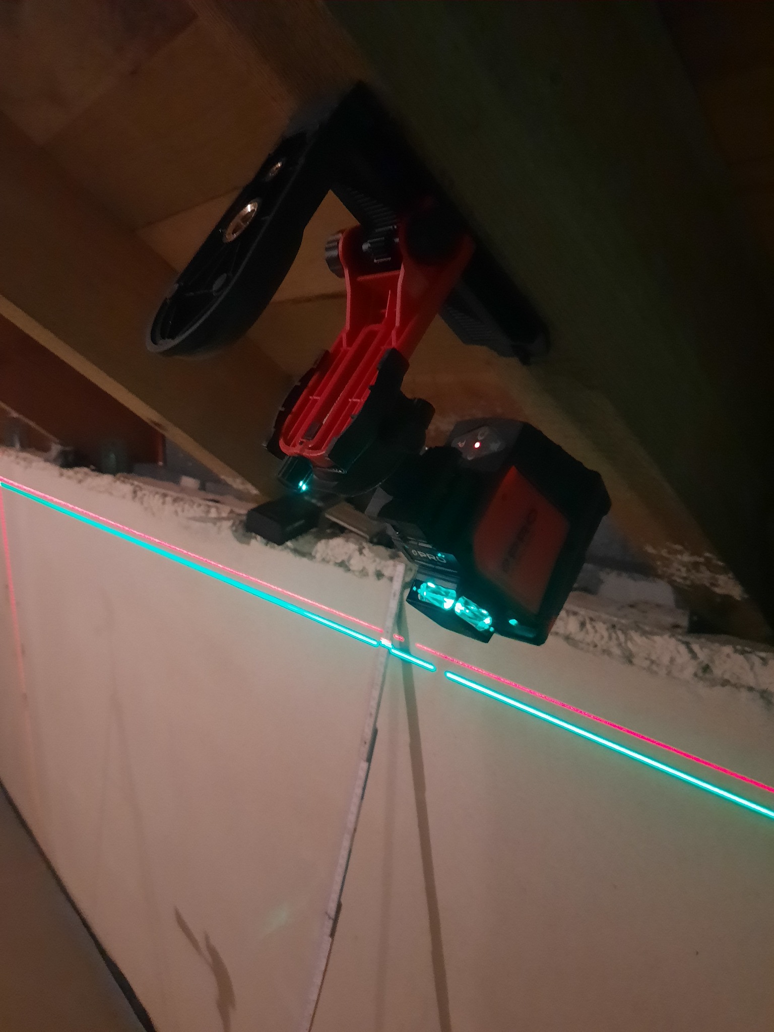 Laser krzyżowy zamontowany na drewnianej konstrukcji poddasza, emitujący zieloną i czerwoną linię na ścianę. Narzędzie do precyzyjnego wyznaczania poziomów.