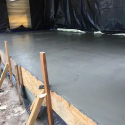 Świeżo wylana, mokra wylewka betonowa na podłożu, zabezpieczona deskowaniem z płyty OSB i folią hydroizolacyjną, widoczne drewniane podpory i fragmenty izolacji termicznej.