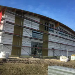 Budynek w trakcie ocieplania, widoczne rusztowania, izolacja z wełny mineralnej, białe bloczki, worki z materiałami budowlanymi i fragmenty blachy na ziemi.