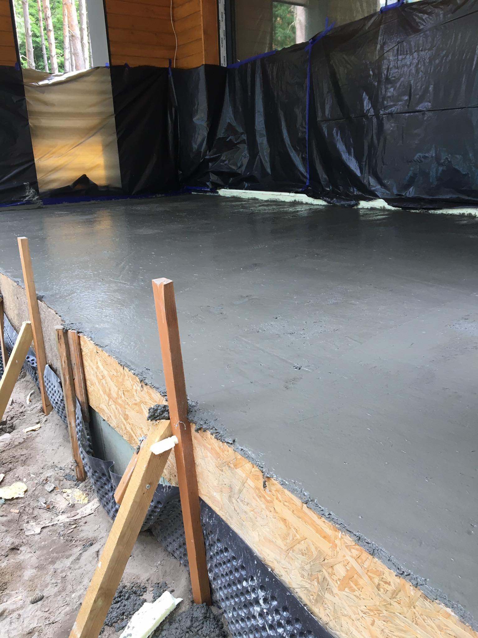 Świeżo wylana, mokra wylewka betonowa na podłożu, zabezpieczona deskowaniem z płyty OSB i folią hydroizolacyjną, widoczne drewniane podpory i fragmenty izolacji termicznej.