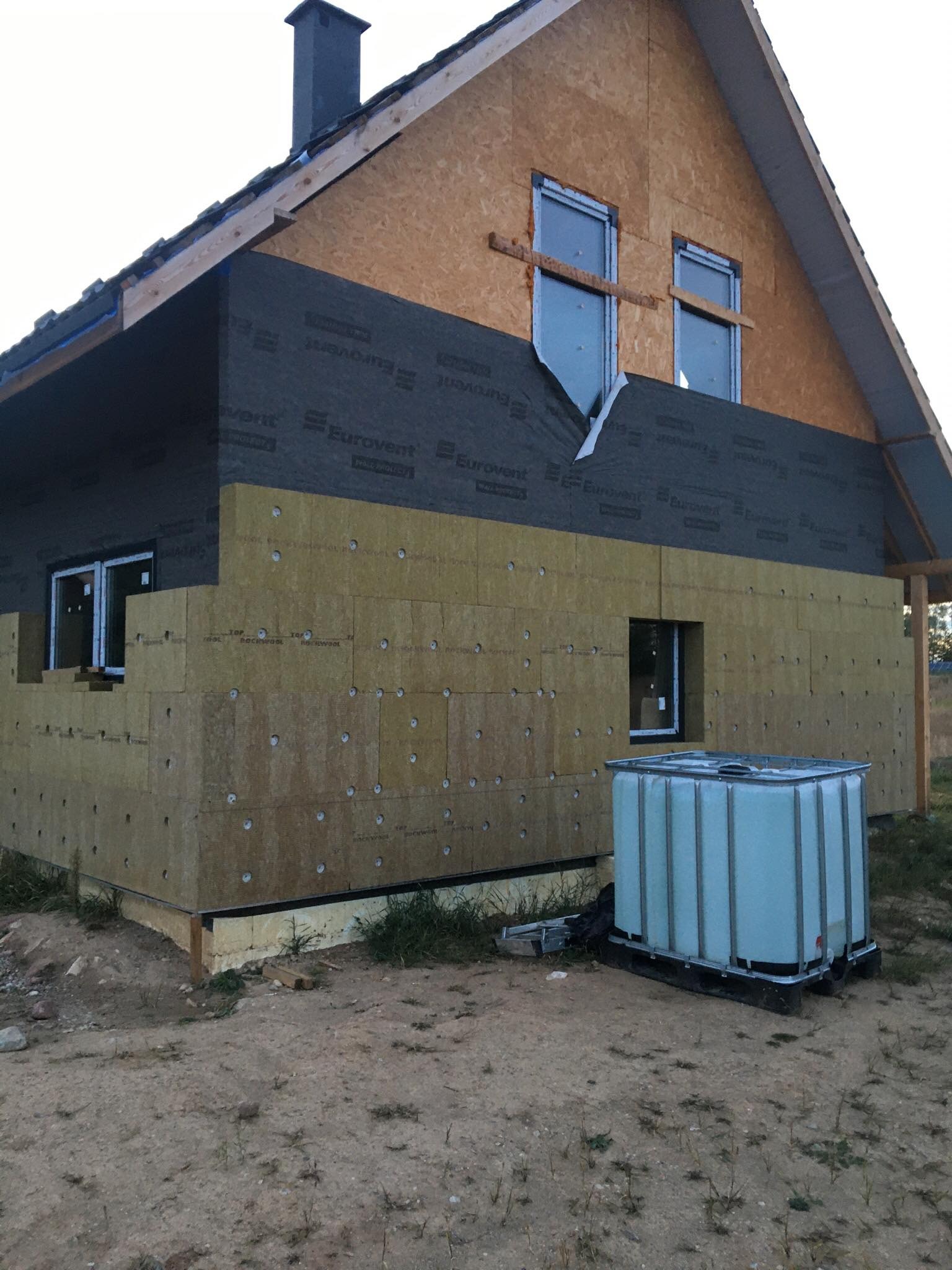 Dom jednorodzinny w trakcie ocieplania, widoczne płyty izolacyjne z wełny mineralnej na ścianie szczytowej, membrana wiatroizolacyjna Eurovent, okna częściowo zakryte folią ochronną, zbiornik...