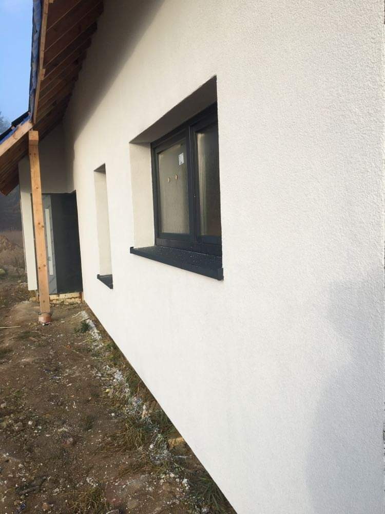 Świeżo otynkowana, biała elewacja domu jednorodzinnego z czarnymi ramami okiennymi, widok z boku, widoczne elementy konstrukcji dachu i fragment terenu z resztkami śniegu.