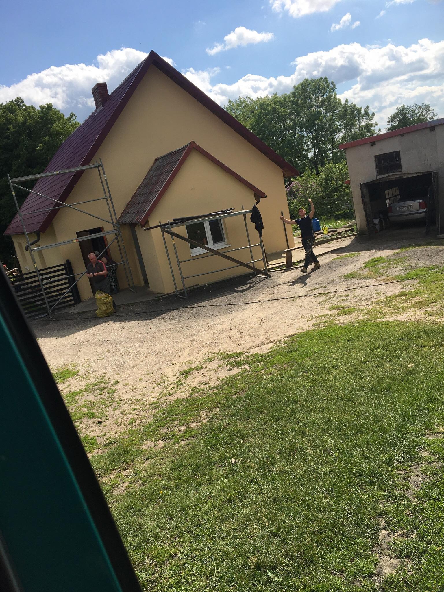 Żółty dom jednorodzinny z ciemnoczerwonym dachem w trakcie prac elewacyjnych, otoczony rusztowaniem; jeden mężczyzna pracuje przy elewacji, drugi stoi obok z podniesionymi rękami w geście radości...