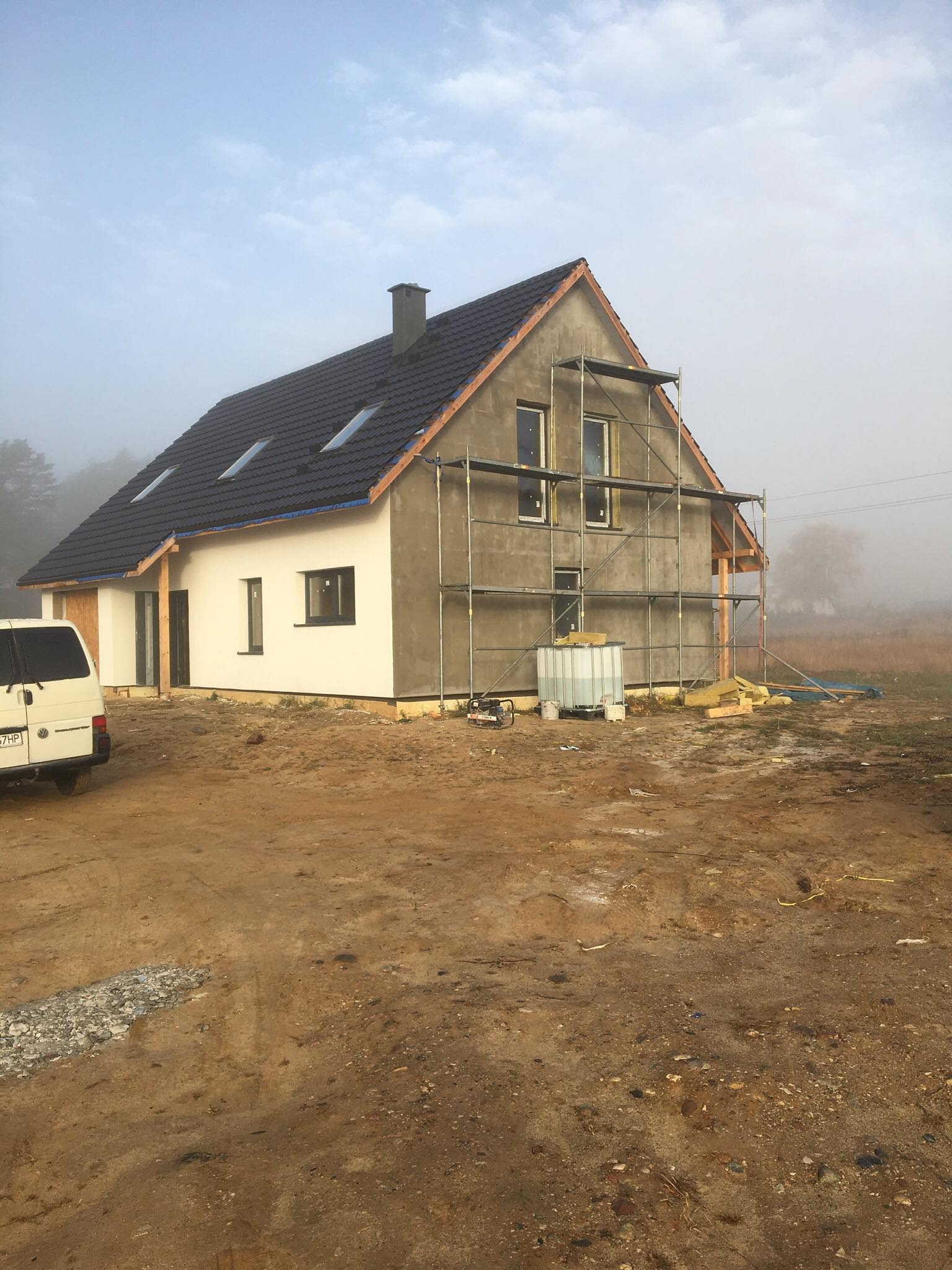 Nowy dom jednorodzinny w trakcie budowy, częściowo otynkowany, z rusztowaniem i widocznym ociepleniem poddasza, w tle mgła.