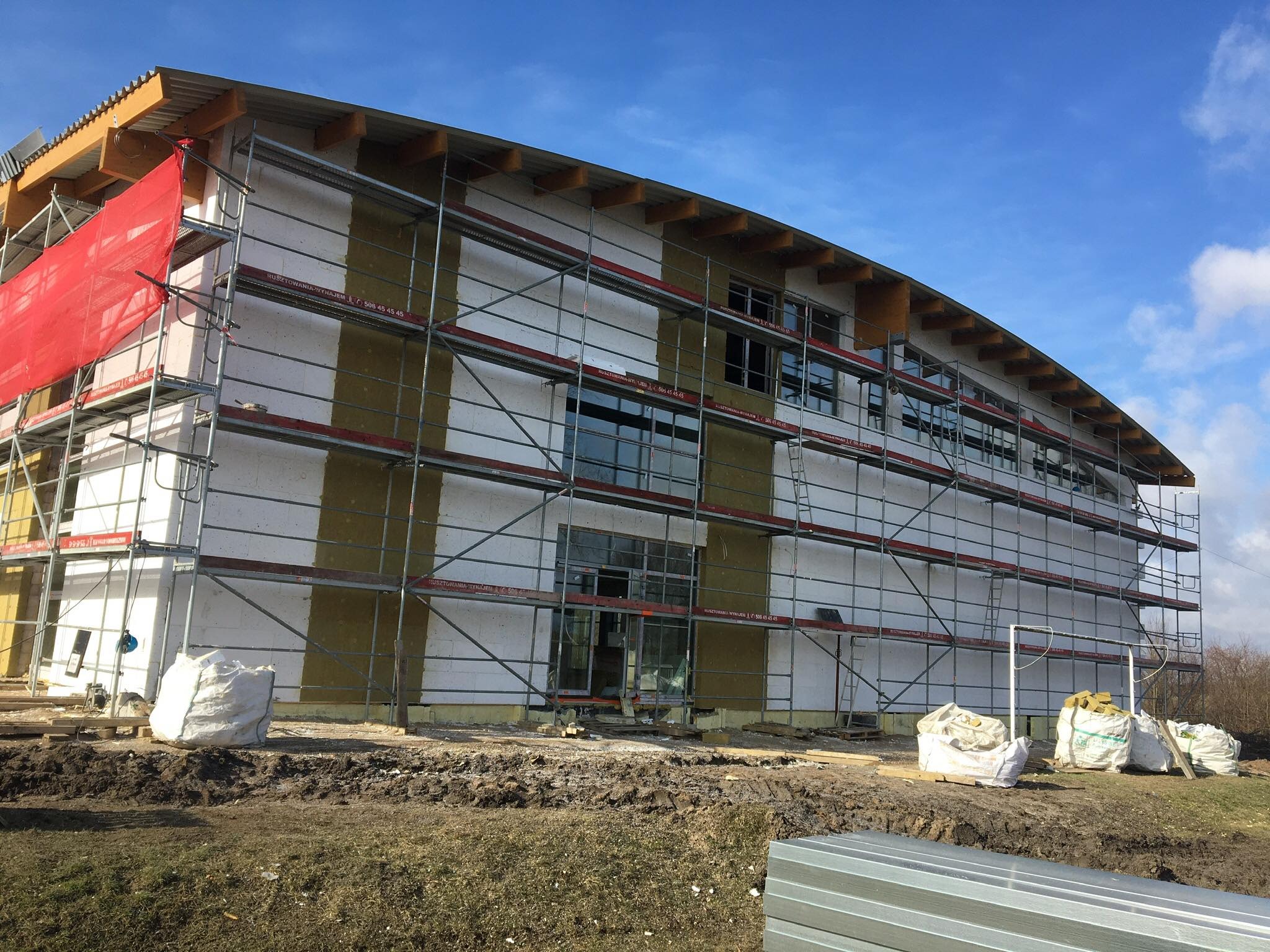 Budynek w trakcie ocieplania, widoczne rusztowania, izolacja z wełny mineralnej, białe bloczki, worki z materiałami budowlanymi i fragmenty blachy na ziemi.