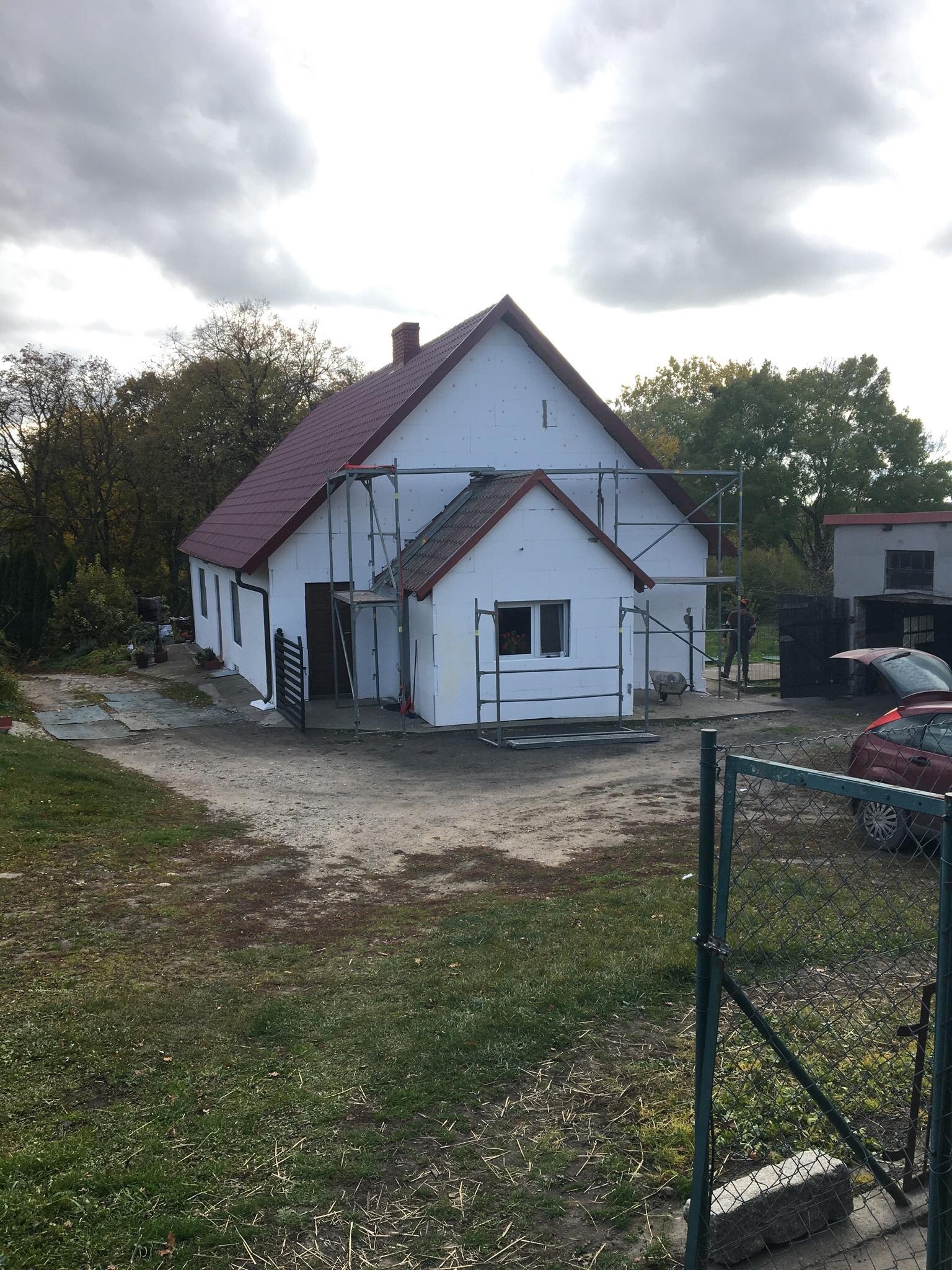 Dom jednorodzinny w trakcie ocieplania styropianem, widoczne rusztowania, czerwony dach i fragment samochodu z otwartym bagażnikiem w tle.