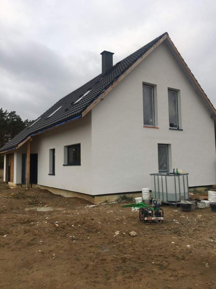 Dom jednorodzinny z białą elewacją w Mrągowie, okna zabezpieczone folią, widoczne narzędzia i pojemniki po farbie, teren wokół domu w trakcie prac wykończeniowych.