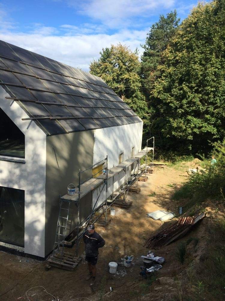 Świeżo otynkowana ściana domu jednorodzinnego z ciemną dachówką, widoczne rusztowania i pracownik w roboczym ubraniu, przygotowany do dalszych prac wykończeniowych.