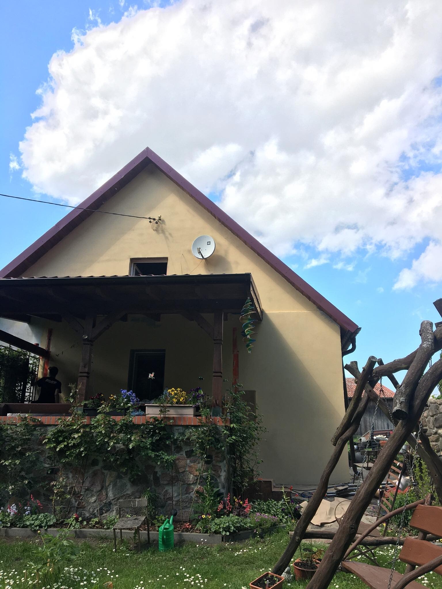 Jasnożółty dom jednorodzinny z brązowym dachem, werandą z kamienną podmurówką i kolorowymi kwiatami w donicach, na elewacji widoczna antena satelitarna.