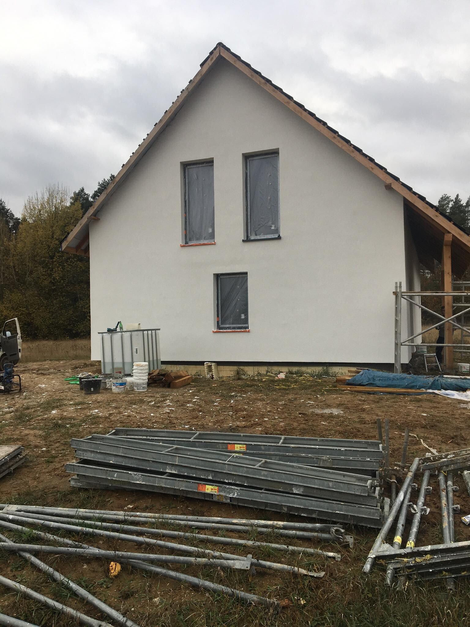 Dom jednorodzinny w trakcie prac elewacyjnych, z oknami zabezpieczonymi folią, widoczne rusztowania, pojemnik na wodę i materiały budowlane na placu budowy.