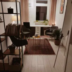 Jasny gabinet z biurkiem, dwoma krzesłami, dywanem orientalnym i regałem z książkami, oświetlony lampą sufitową i lampką biurkową, z elementami dekoracyjnymi w postaci roślin doniczkowych i obrazu...