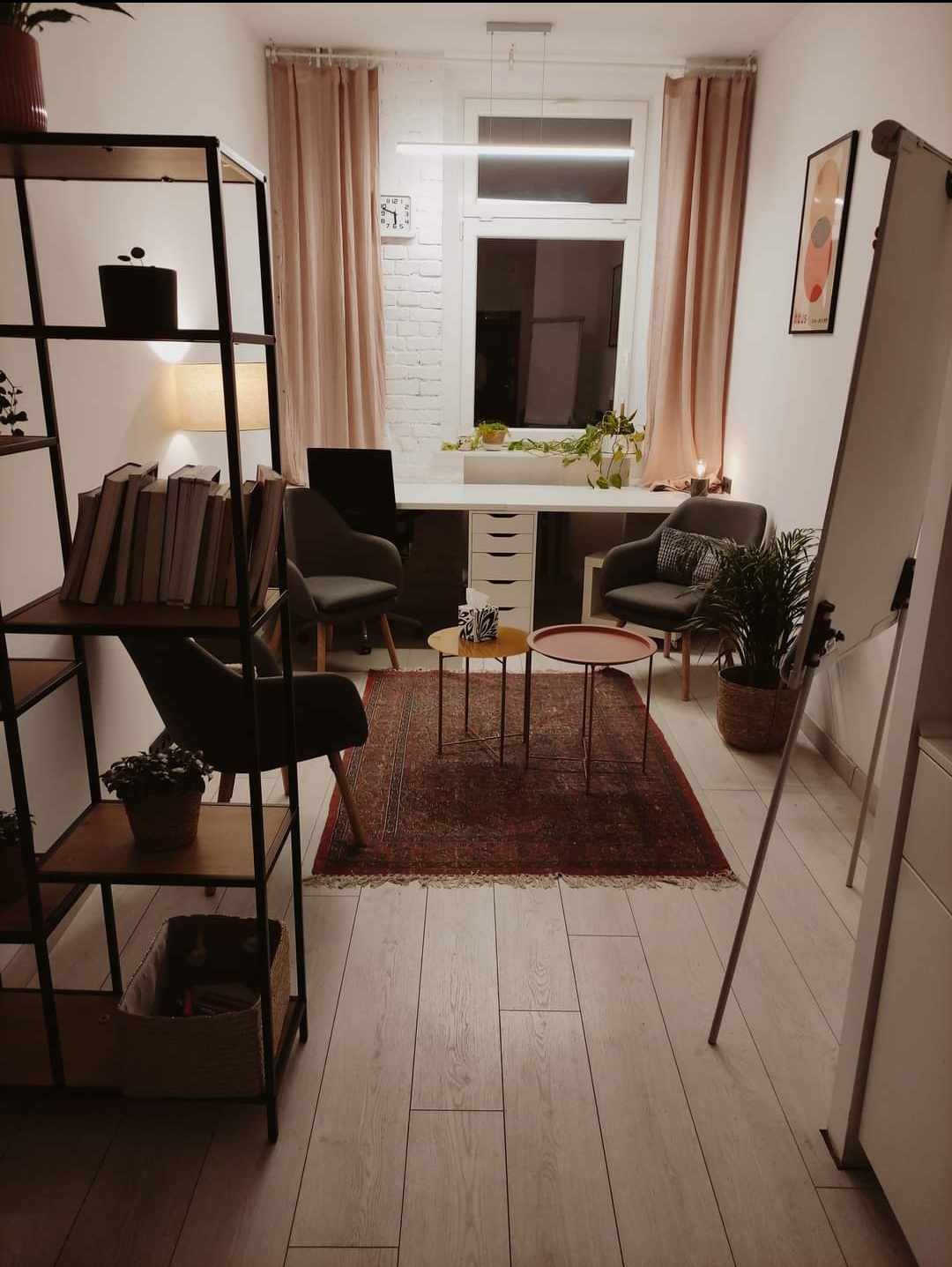 Jasny gabinet z biurkiem, dwoma krzesłami, dywanem orientalnym i regałem z książkami, oświetlony lampą sufitową i lampką biurkową, z elementami dekoracyjnymi w postaci roślin doniczkowych i obrazu...